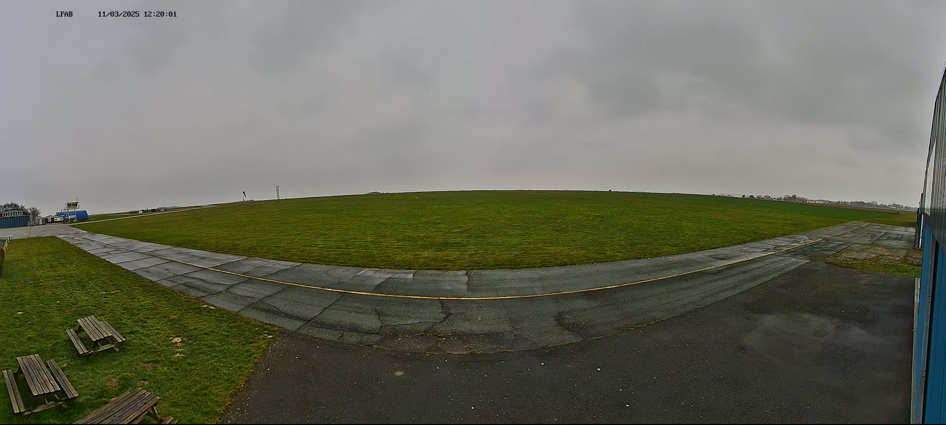 A grassy airfield with a paved taxiway curves towards a small building under an overcast sky.