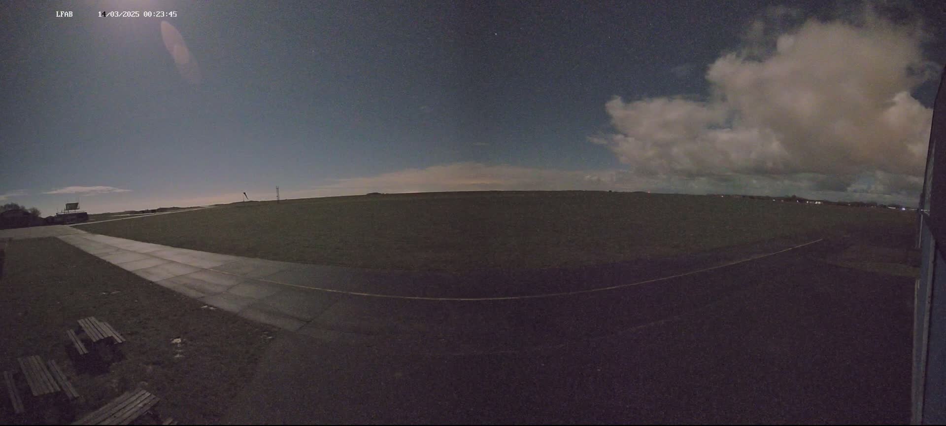 A paved path extends from the bottom of the frame toward a flat, dark field under a night sky with partly cloudy conditions.