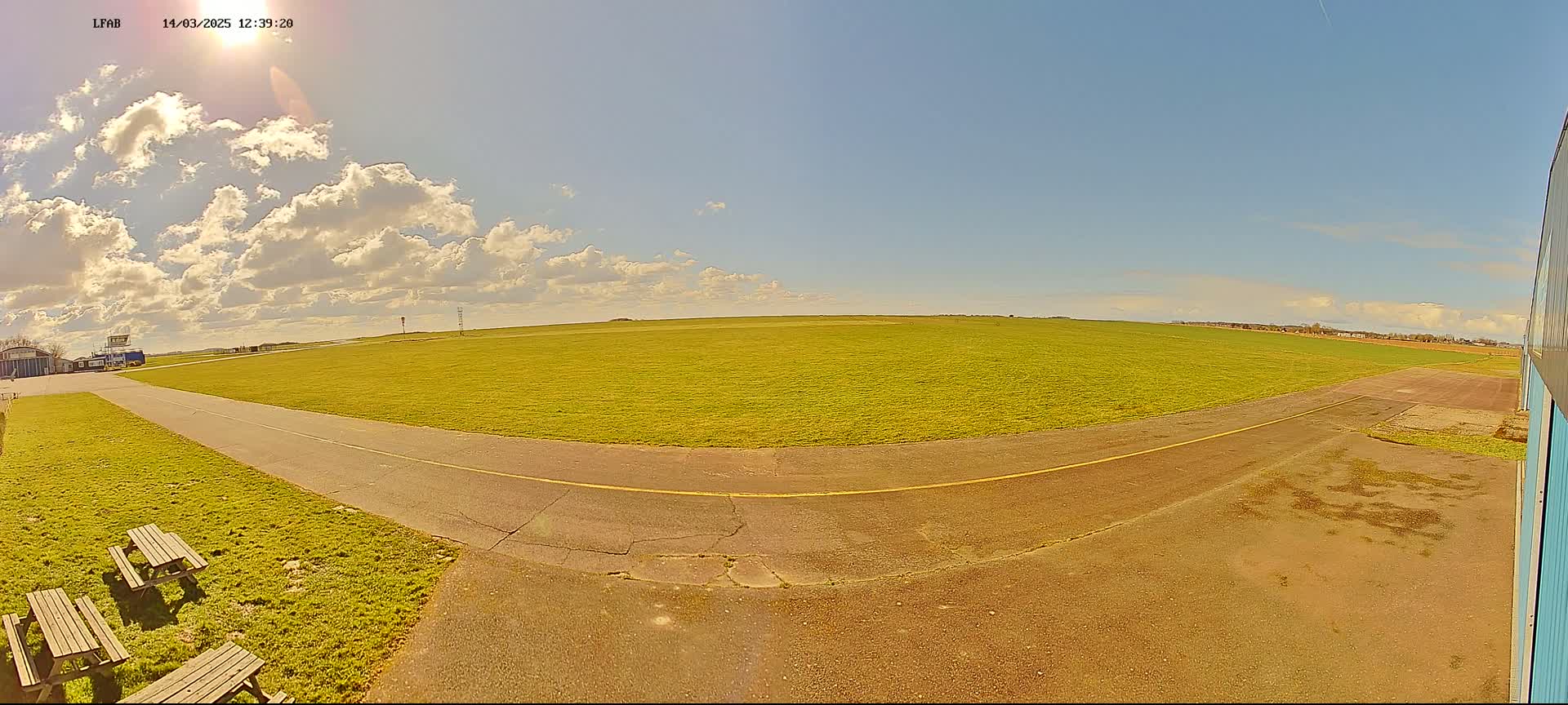 A sunny day with scattered clouds reveals a grassy airfield with a paved taxiway, picnic tables, and a portion of a hangar.