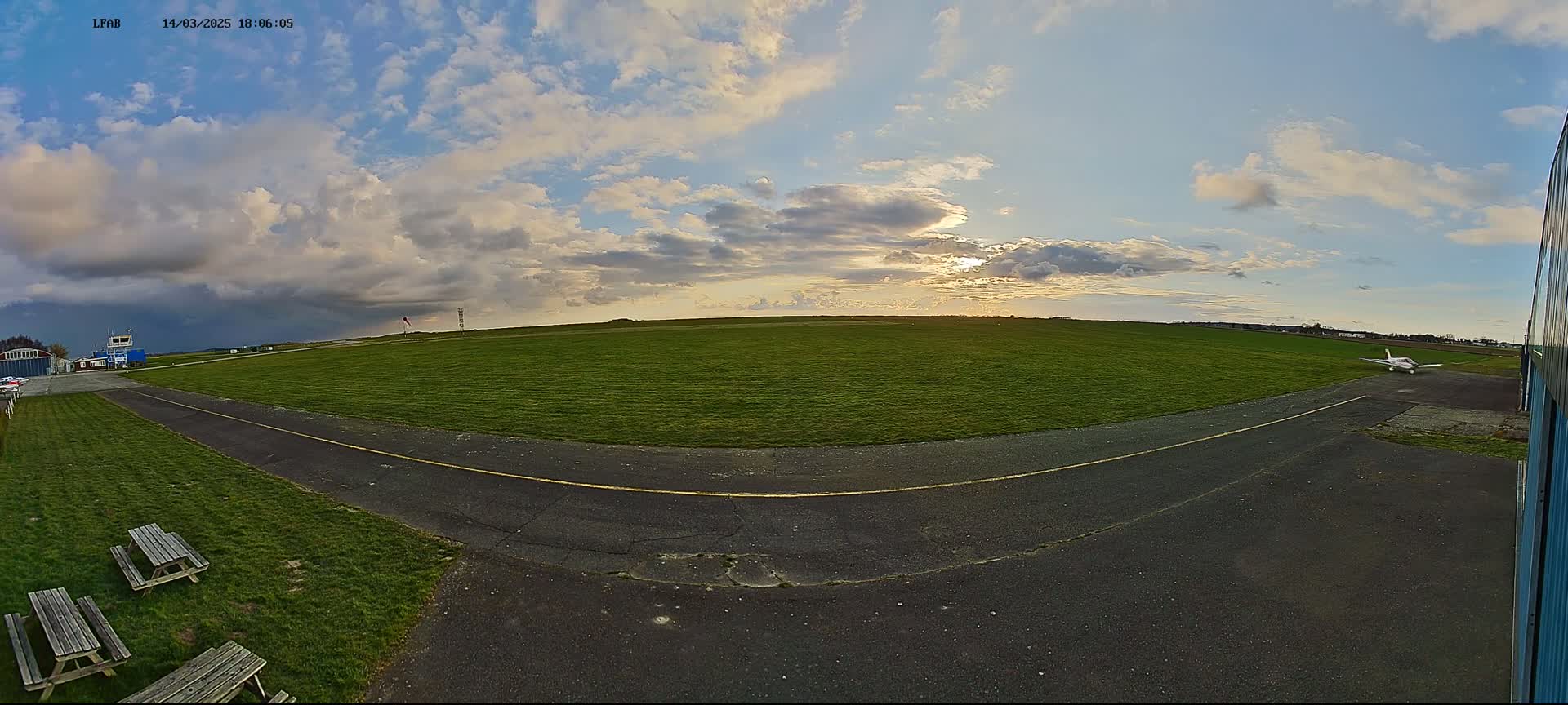 A small airplane sits on a grassy airfield under a partly cloudy sky with the sun partially visible.