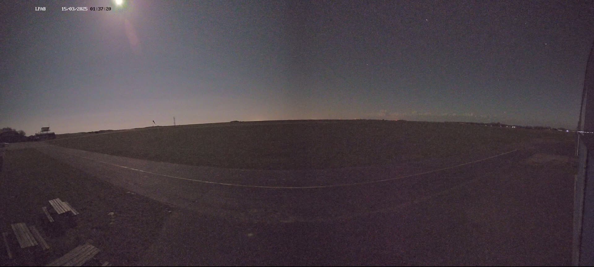 A nighttime, low-light view of a flat, dark landscape with a paved road and a few park benches under a mostly clear, dark sky showing a bright moon.