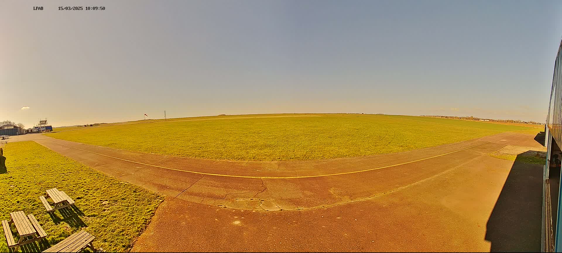 A sunny airfield with a grassy field, a paved taxiway, and picnic benches is seen from a low vantage point.