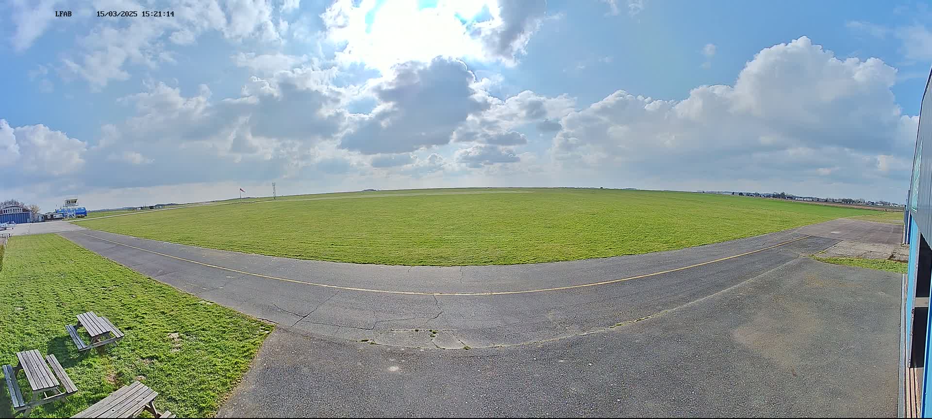 A grassy airfield under a partly sunny sky with scattered cumulus clouds is seen from a ground-level perspective, showing a paved taxiway curving through the grass.