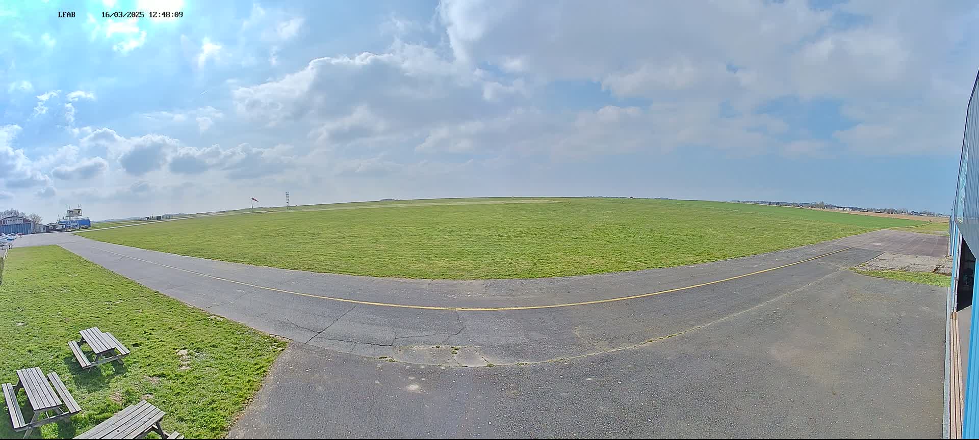 A grassy airfield with a paved taxiway and picnic tables under a partly cloudy sky.