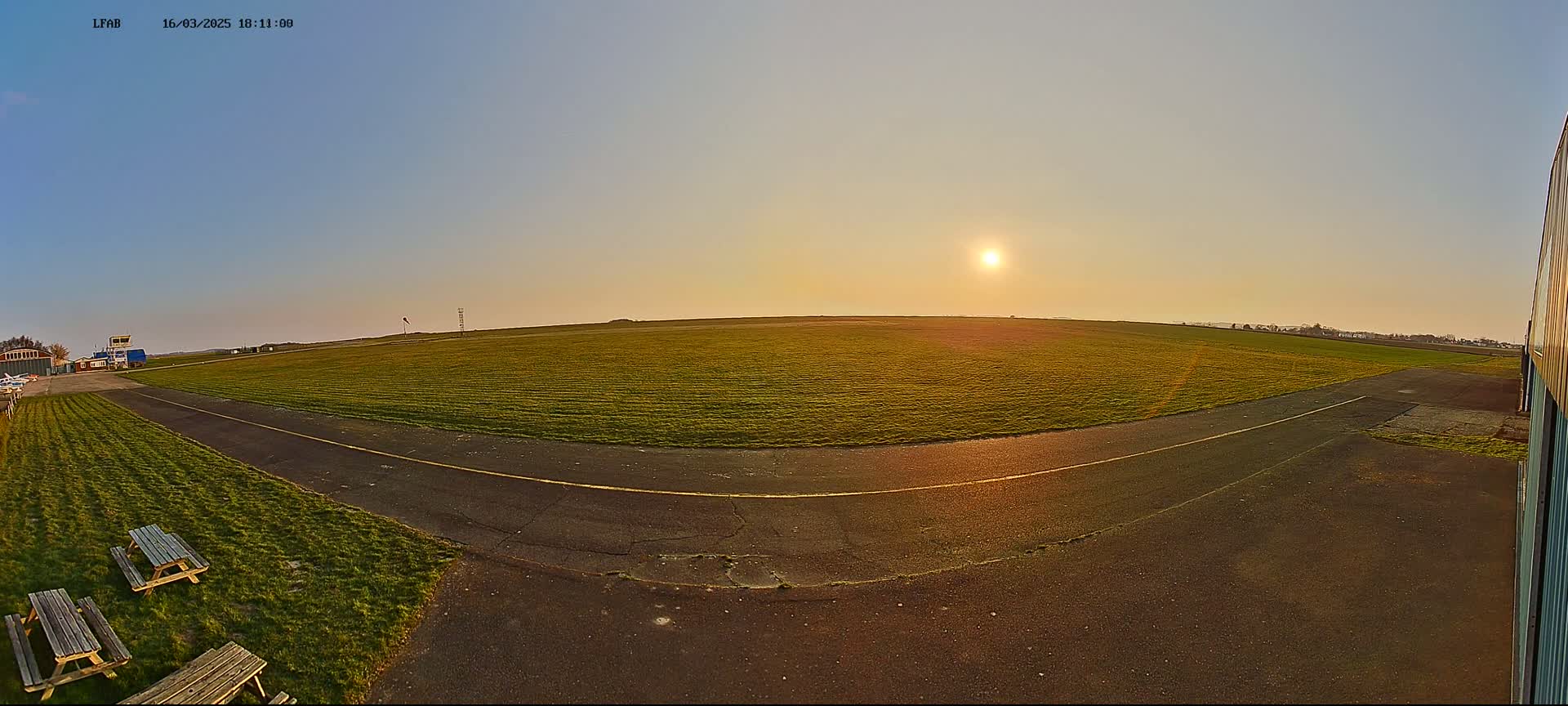 A grassy airfield with a paved taxiway is shown at sunset under a clear sky.
