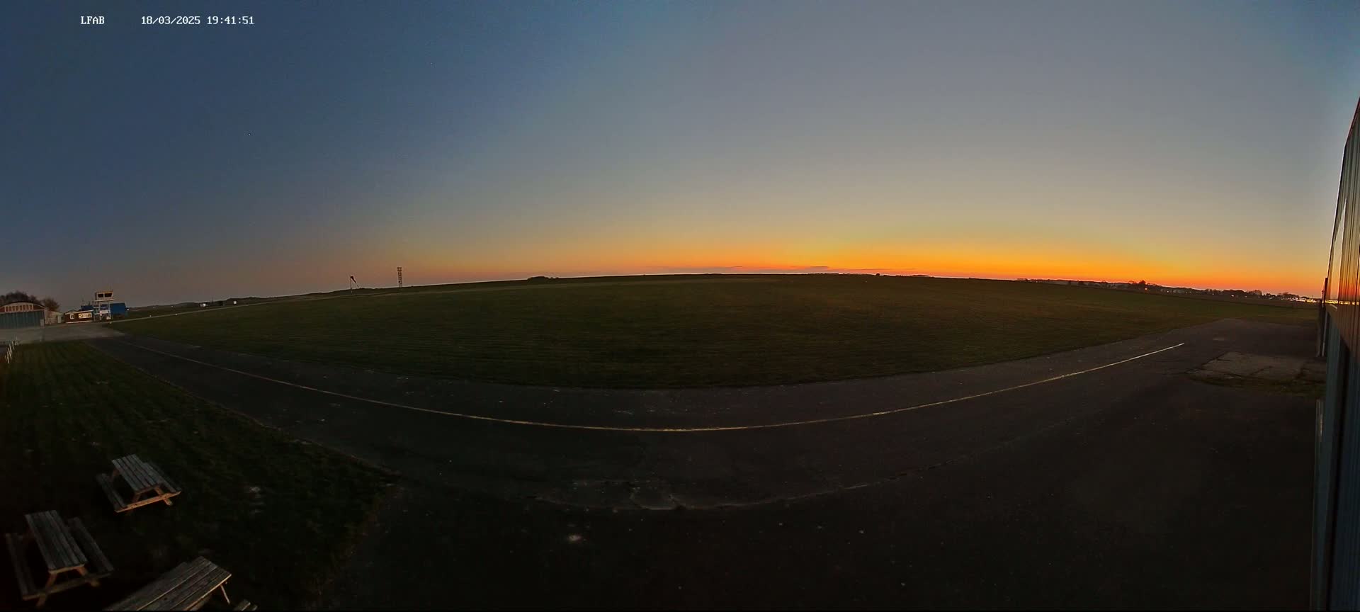 A grassy airfield at sunset, with a paved taxiway and picnic tables visible near a hangar.