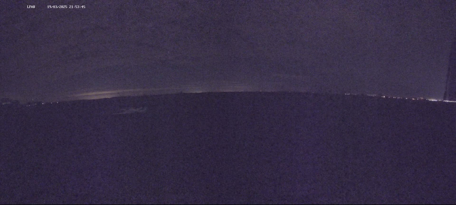 A dark, nighttime landscape is visible, showing a flat horizon with faint lights in the distance under a mostly clear sky.