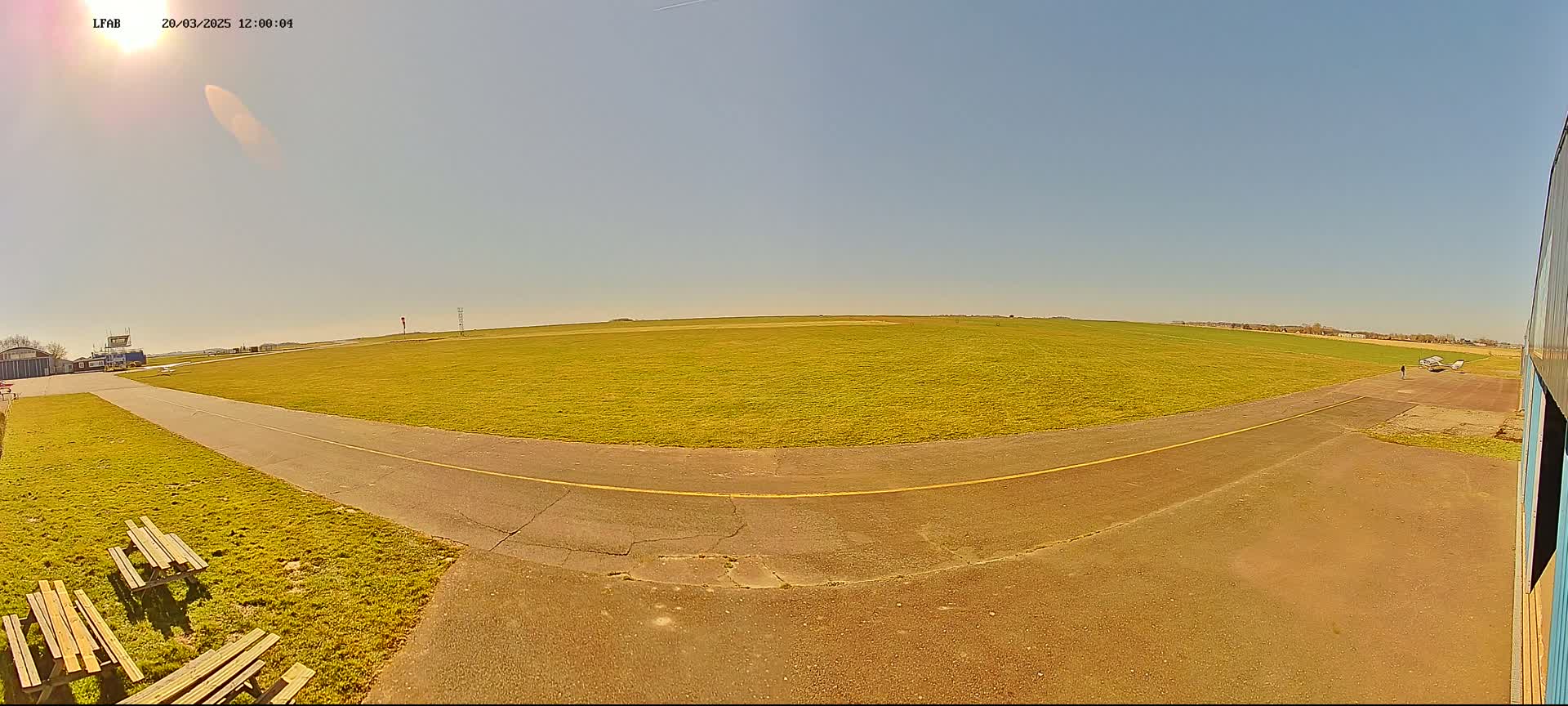 A sunny day at a small airfield shows a grassy field with a paved taxiway and picnic tables near a hangar.