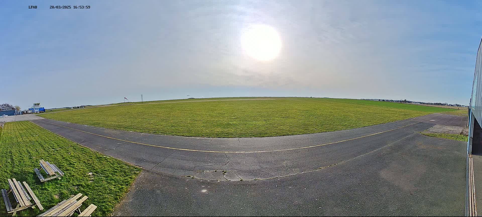 A sunny day reveals a grassy airfield with a paved taxiway, bordered by picnic tables and distant buildings.