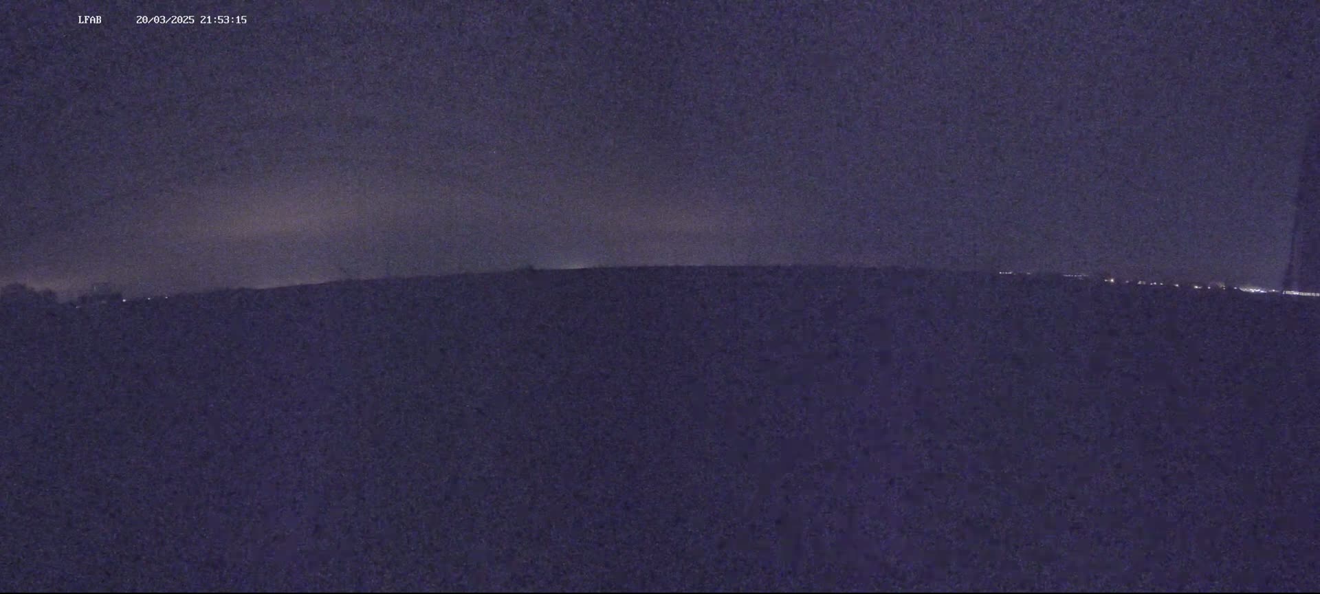 A dark, nighttime landscape shows a flat horizon with a few distant lights visible under a mostly clear, dark sky.