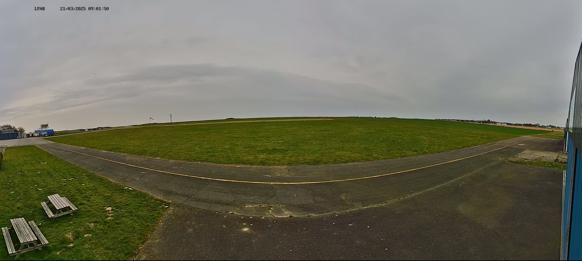 A grassy airfield with a paved taxiway and picnic tables is visible under an overcast sky.