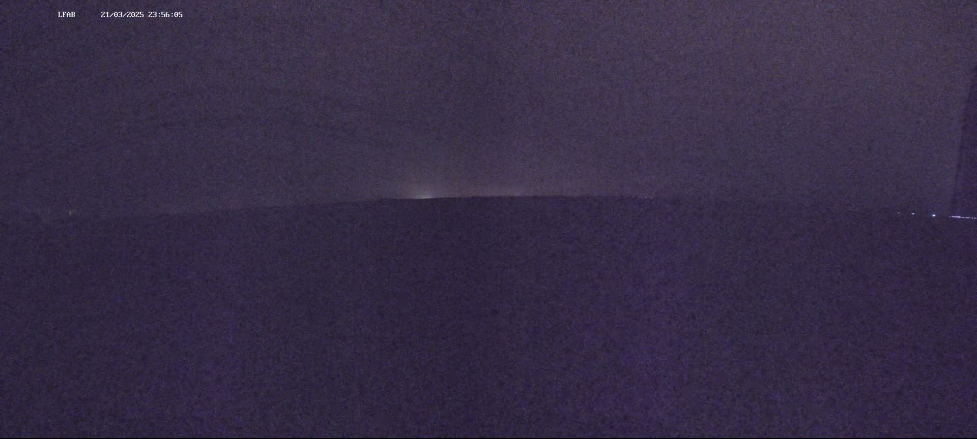 A dark, nighttime landscape shows a distant, faintly lit horizon under a mostly cloudy sky.
