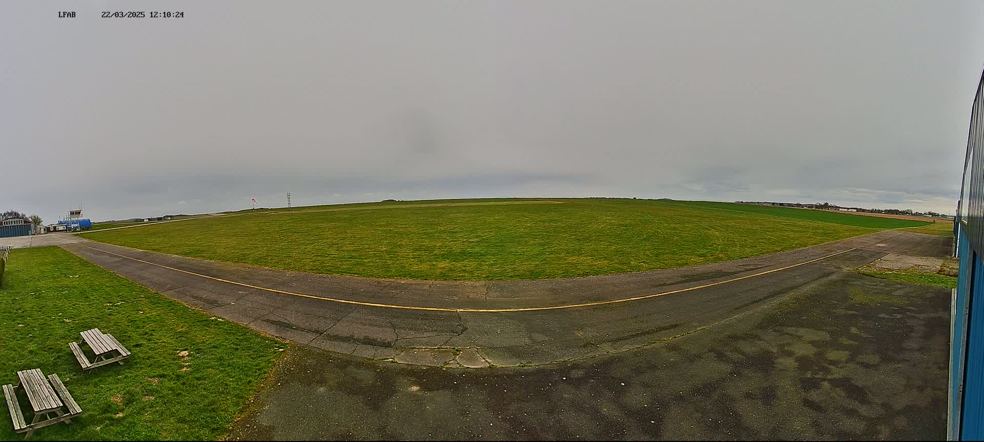 A grassy airfield under an overcast sky is seen from a ground-level perspective, featuring a paved taxiway and a couple of picnic benches.