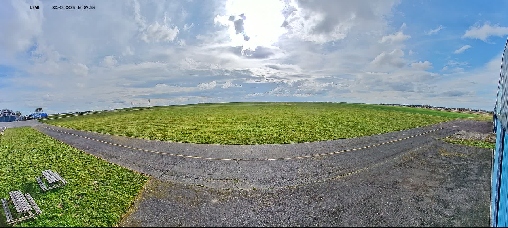 A wide, grassy airfield under a partly sunny sky with scattered clouds is shown, featuring a paved taxiway curving towards a hangar.