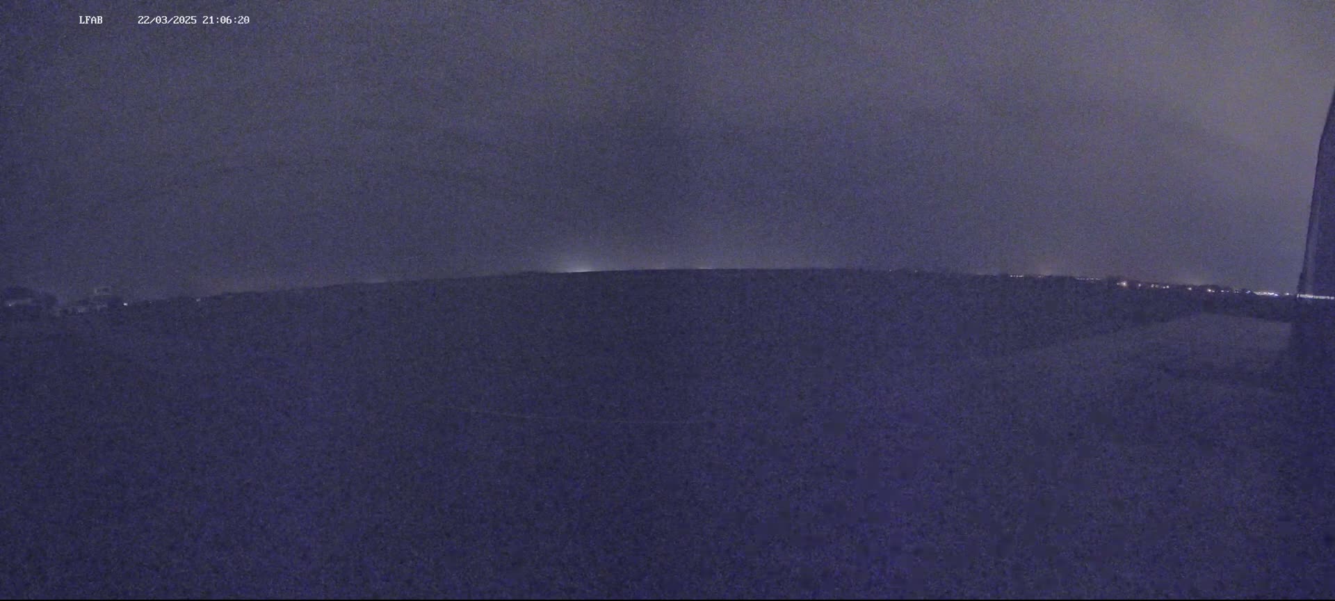 A dark, nighttime view shows a flat, dark landscape with a faint horizon line and distant lights under a cloudy sky.
