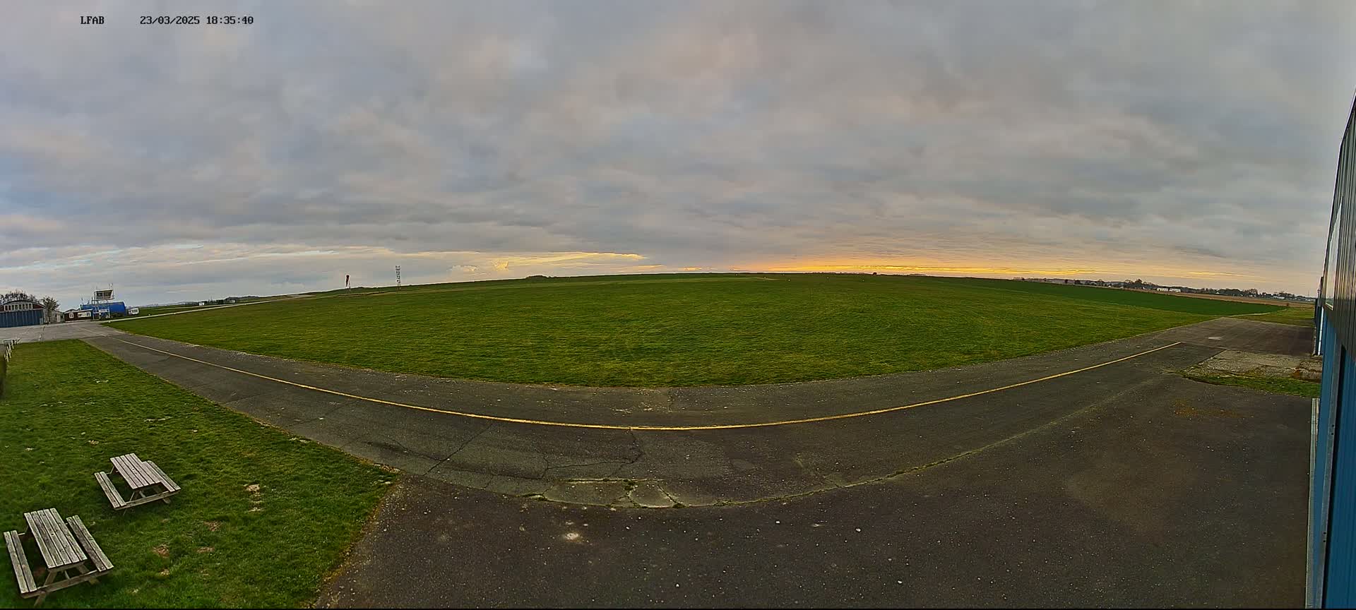 A grassy airfield with a paved taxiway is visible under a cloudy sky with a hint of sunset.