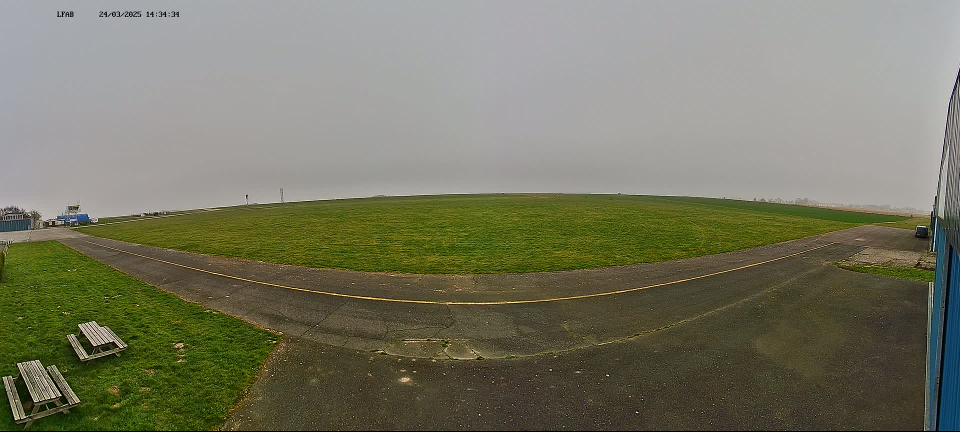 A grassy airfield with a paved runway and picnic tables is visible under an overcast sky.