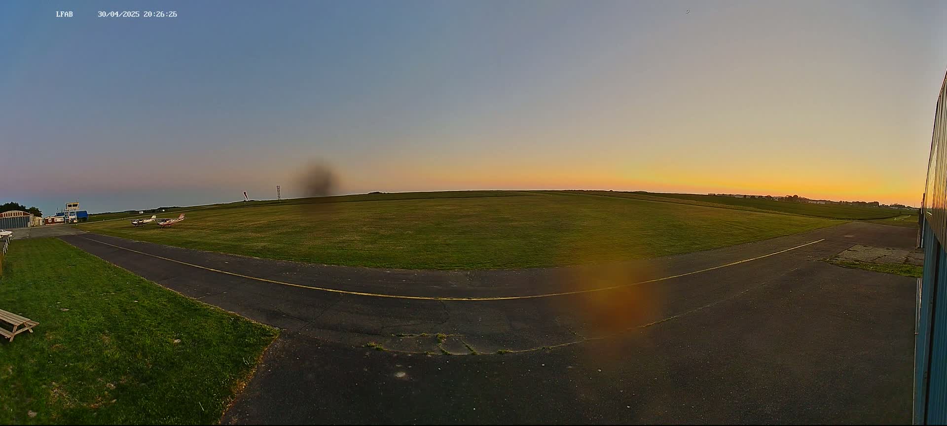 A grassy airfield at sunset, with two small airplanes parked on the tarmac and a paved taxiway leading to a hangar.