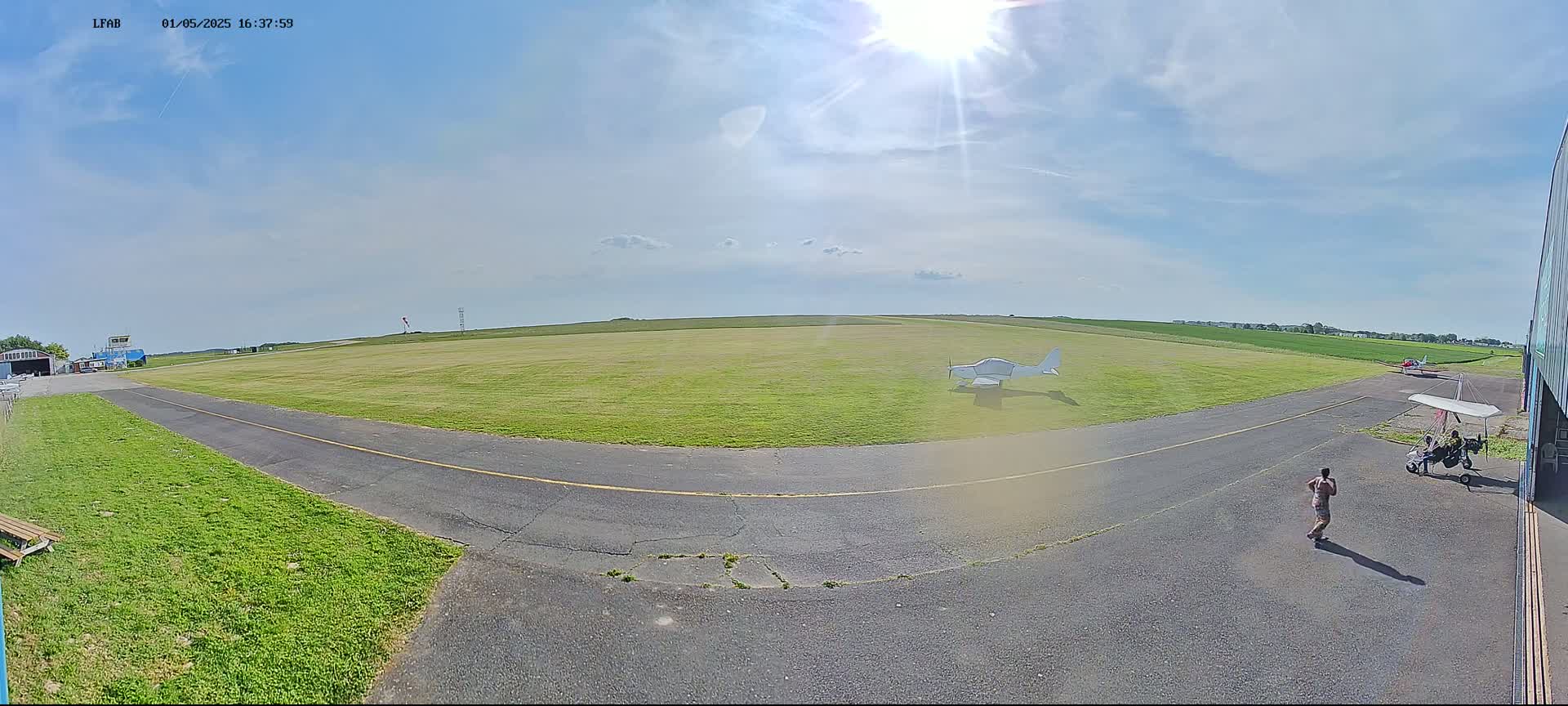 A sunny day at a small airfield shows a grassy field with a small white airplane, a paved taxiway, and a person walking near a hangar.