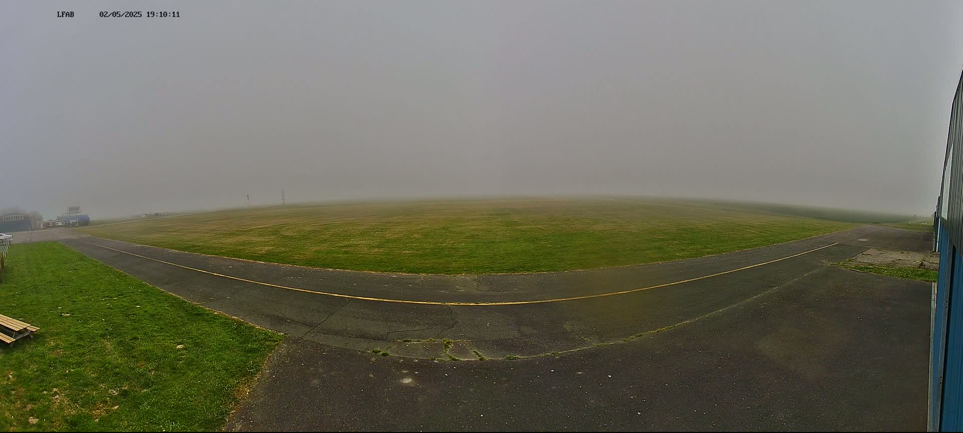 A grassy airfield is shrouded in thick fog, with a paved taxiway curving towards a hangar.