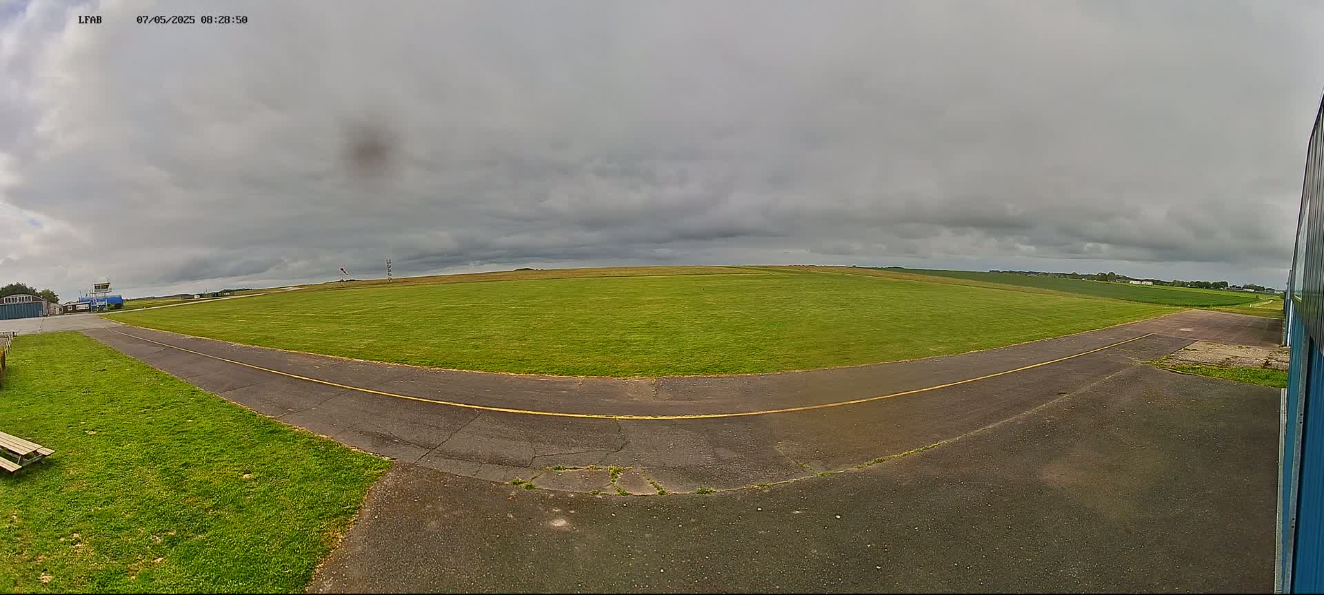 A grassy airfield under a cloudy sky shows a paved taxiway curving towards a hangar.