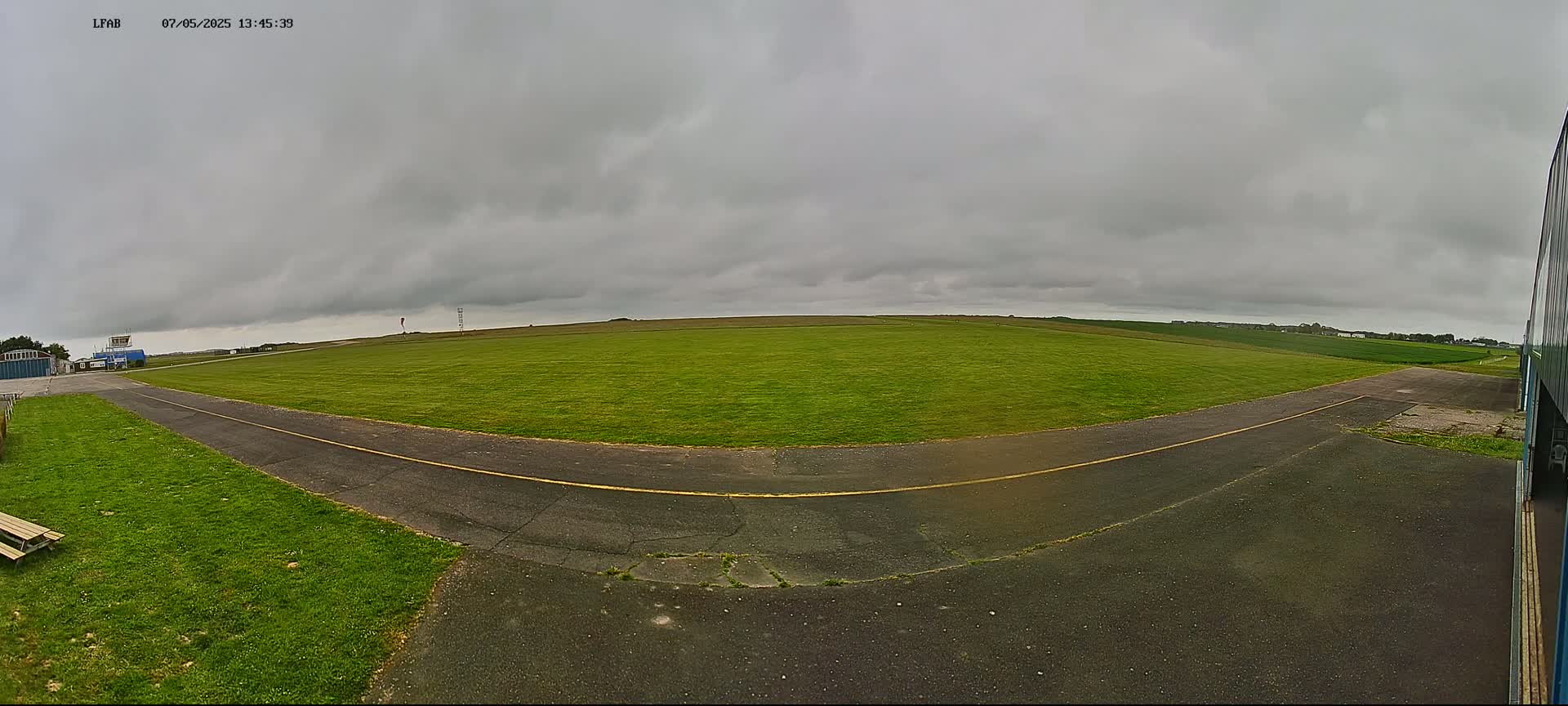 A wide, grassy airfield under an overcast sky is seen from a ground-level perspective, with a paved taxiway curving towards a hangar in the foreground.