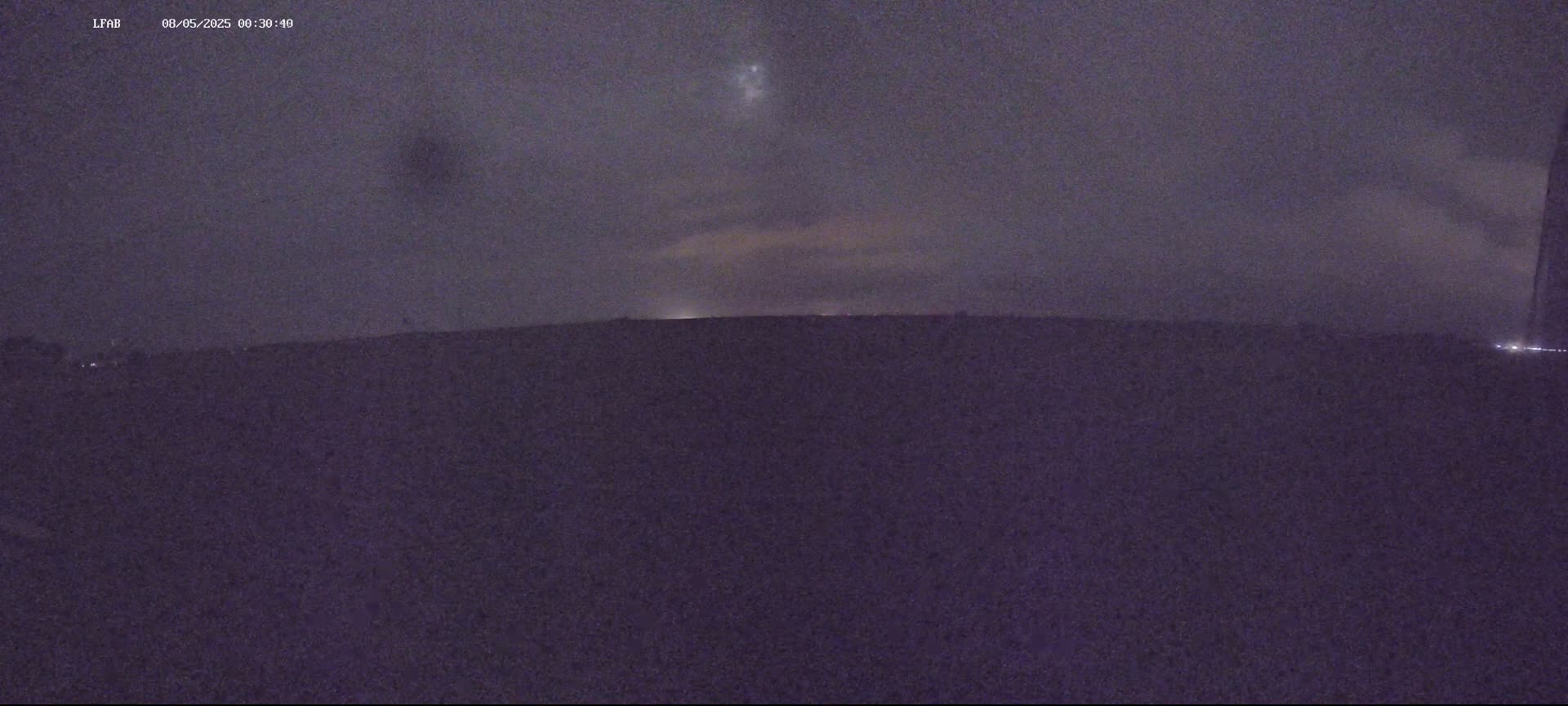A dark, nighttime landscape shows a flat horizon under a mostly cloudy sky with a few faint lights visible in the distance.