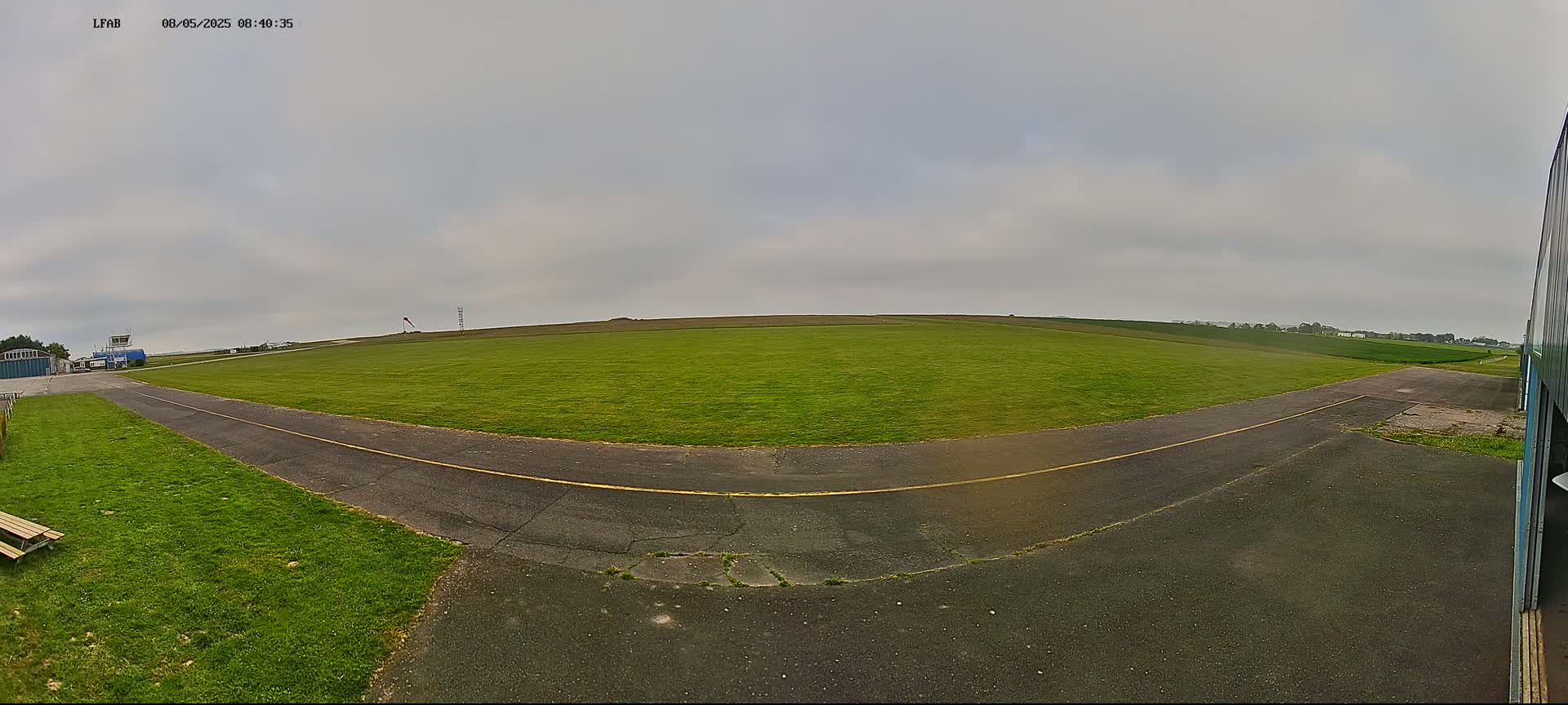A grassy airfield under an overcast sky shows a paved taxiway curving away from a hangar.