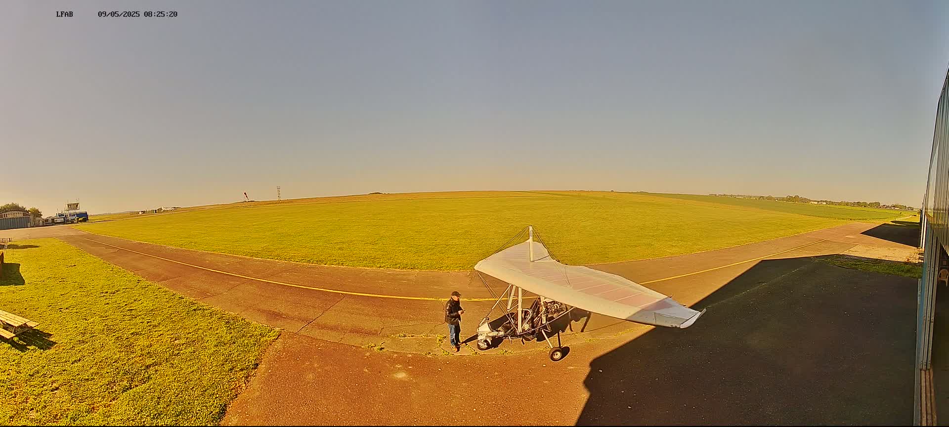 A person stands near a hang glider on a grassy airfield under a clear, sunny sky.