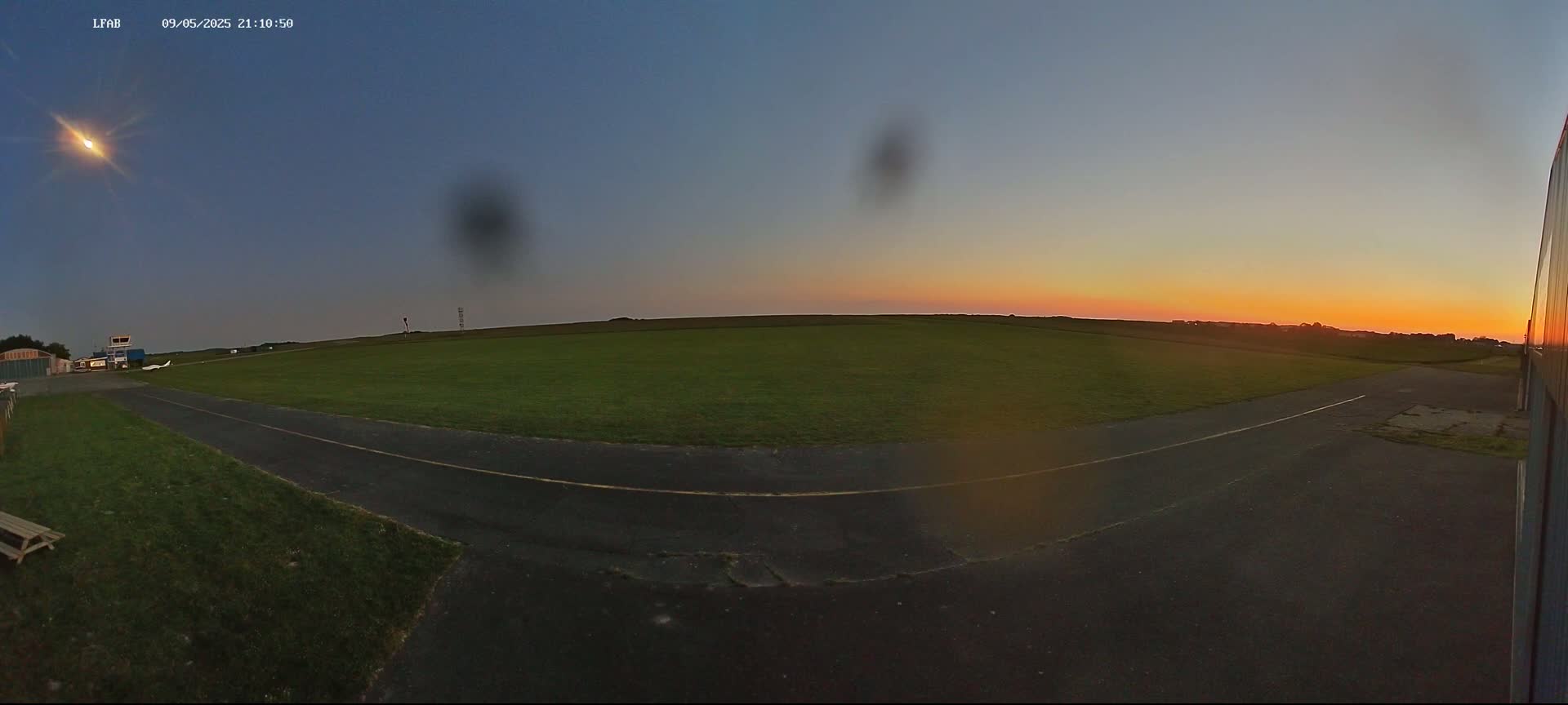 A grassy airfield with a paved taxiway is visible during a sunset with clear skies.