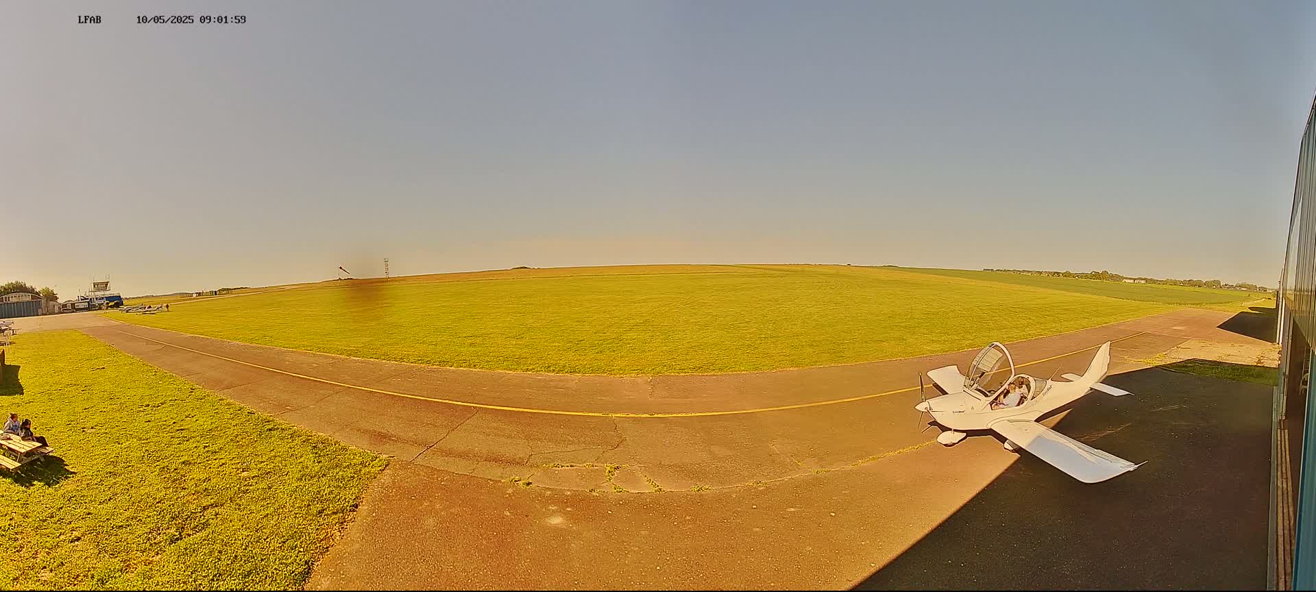A small, white airplane sits on a grassy airfield under a clear, sunny sky.