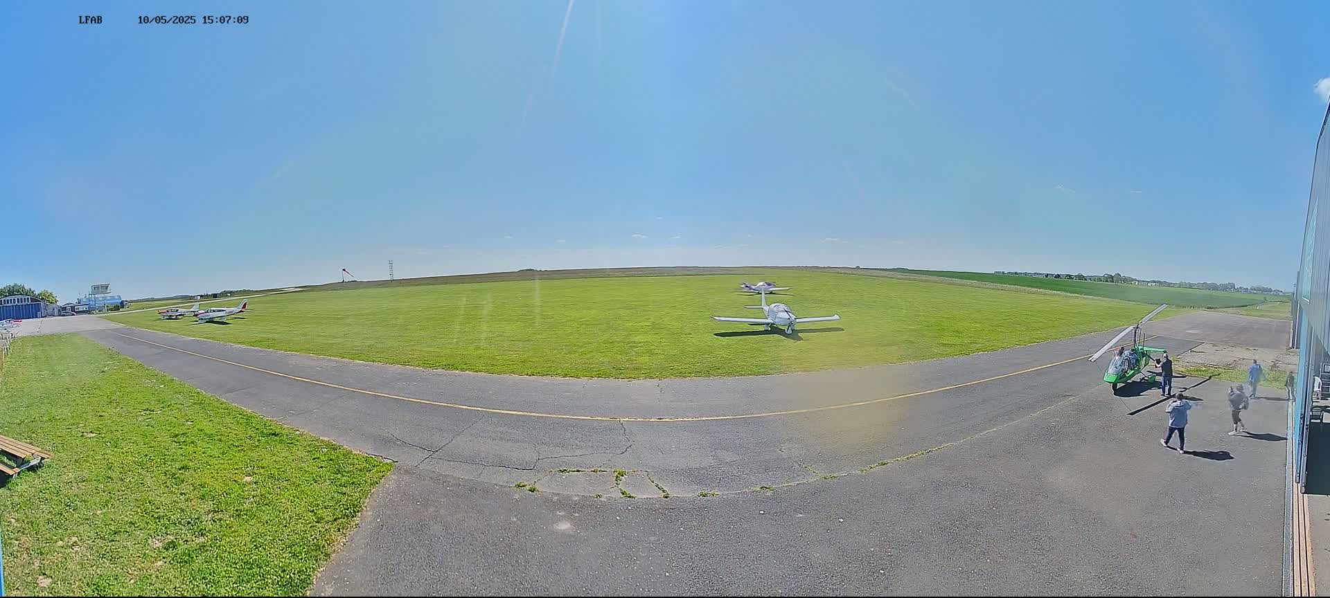 A grassy airfield under a clear blue sky features several small airplanes and a gyrocopter on the tarmac, with a few people standing nearby.