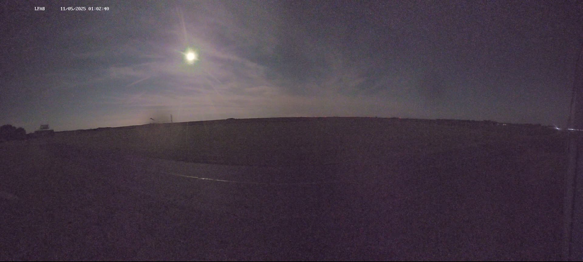A bright moon shines in a mostly clear night sky over a dark, flat landscape.