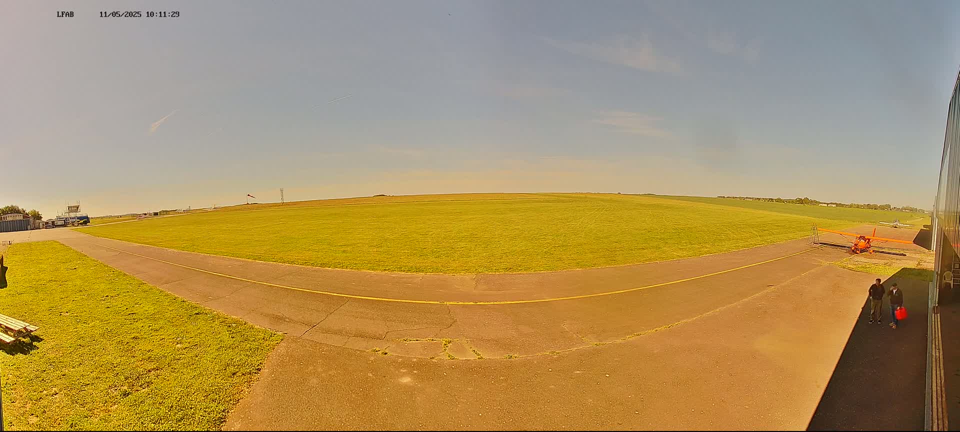 A small orange airplane sits on a grassy airfield under a mostly clear, sunny sky.