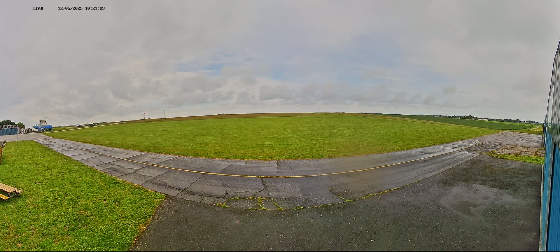 A grassy airfield with a paved taxiway curves to the left under a cloudy sky.