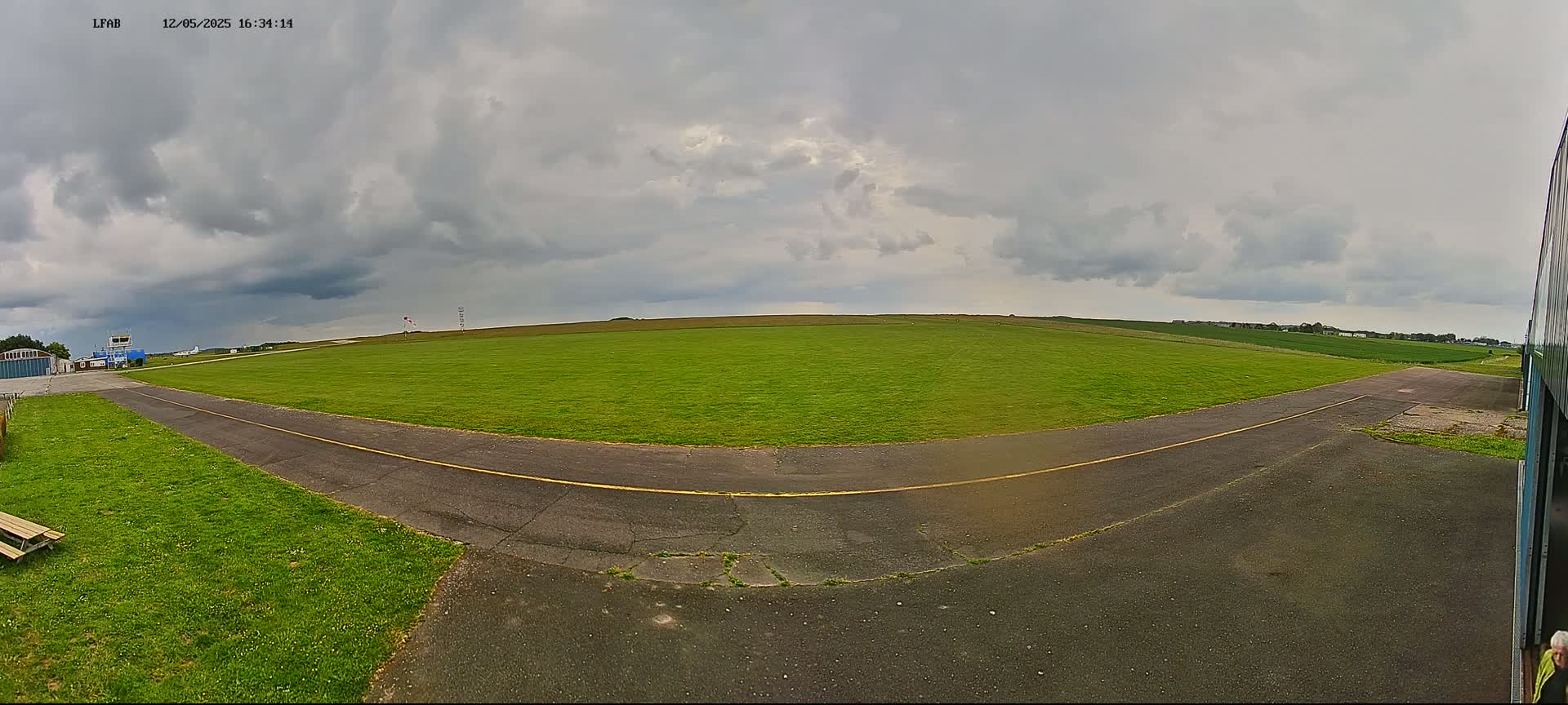 A grassy airfield with a paved taxiway stretches out under a mostly cloudy sky.
