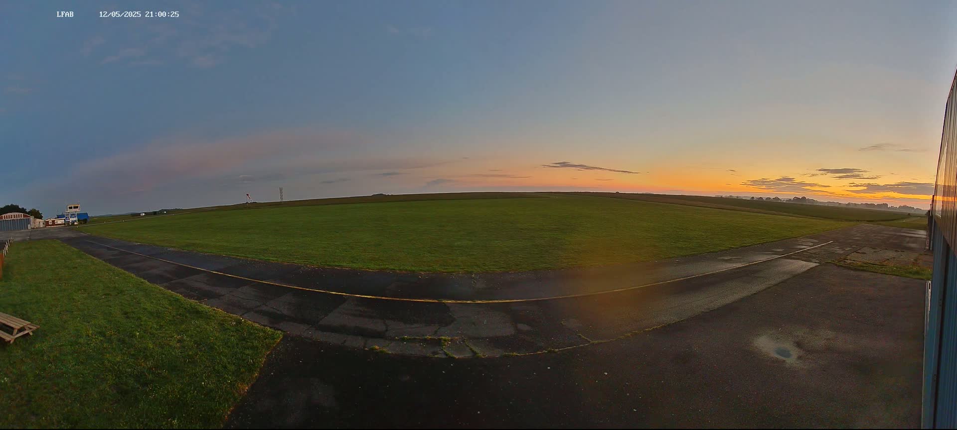 A grassy airfield at sunrise with a paved taxiway, showing a partly cloudy sky with orange and yellow hues at the horizon.