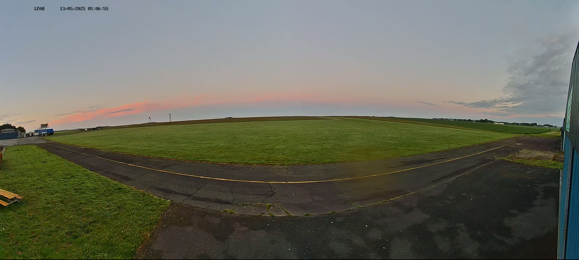 A grassy airfield at sunrise under a mostly clear sky shows a paved taxiway leading to a runway with a small building visible in the distance.