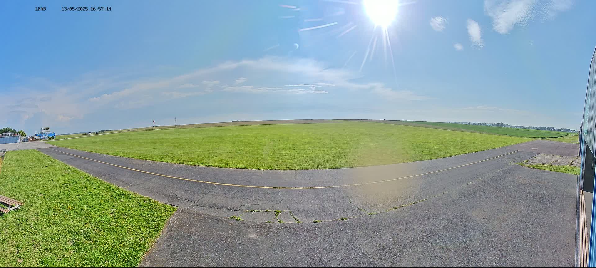 A grassy airfield with a paved taxiway is visible under a mostly sunny sky with a few scattered clouds.