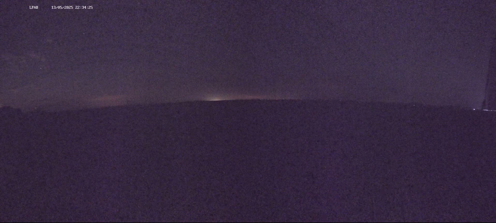 A dark, nighttime landscape shows a dimly lit horizon under a mostly clear, dark purple sky.