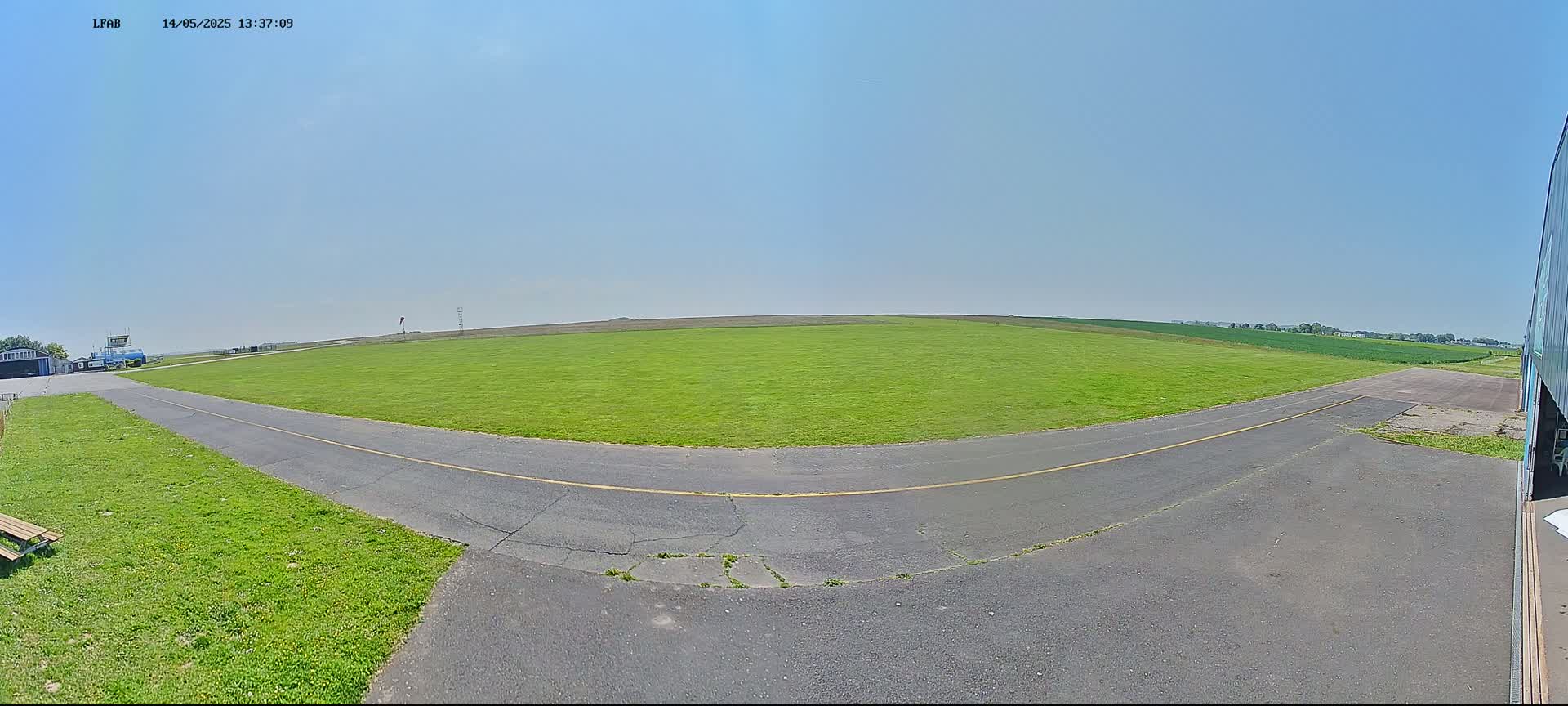A grassy airfield with a paved taxiway curves to the right under a clear, sunny sky.
