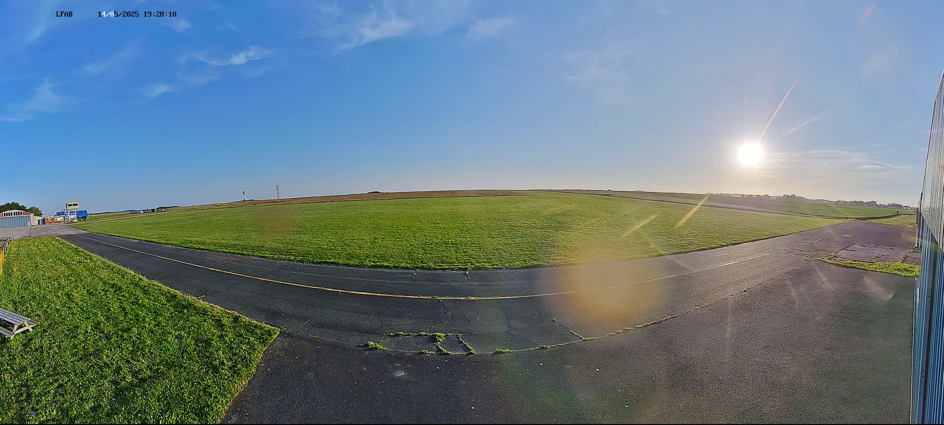 A wide, grassy airfield is bathed in bright sunlight under a mostly clear blue sky, with a paved taxiway leading towards a small control tower.
