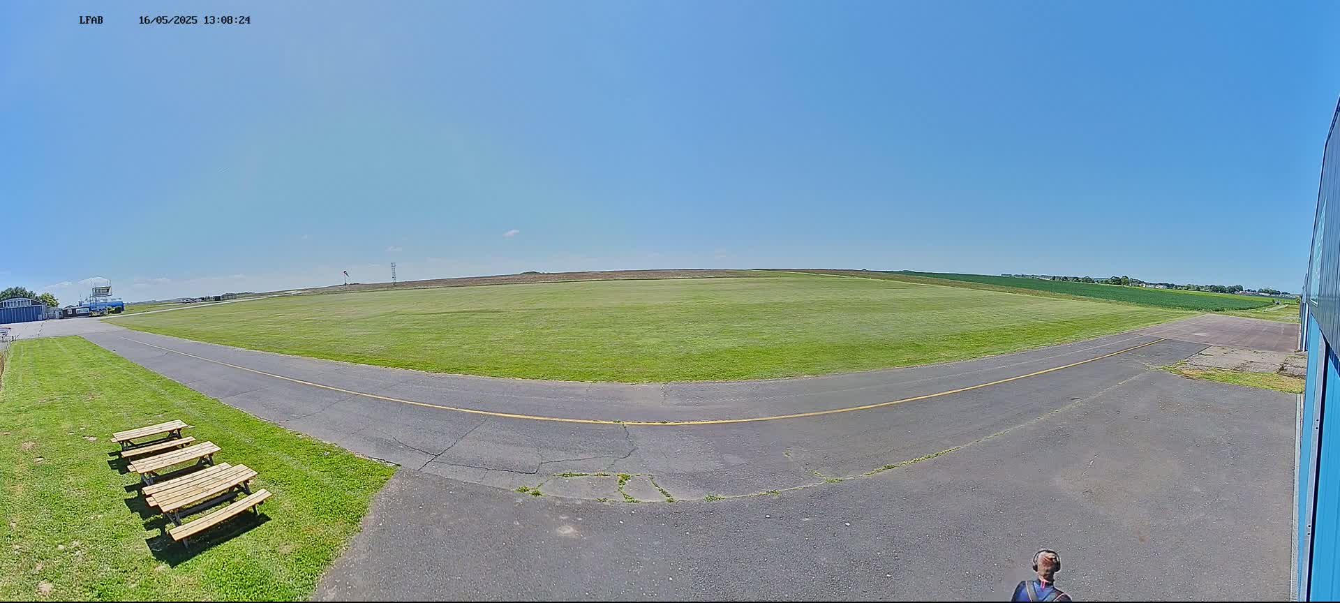 A grassy airfield under a clear blue sky, with picnic tables and a paved taxiway leading to a hangar.