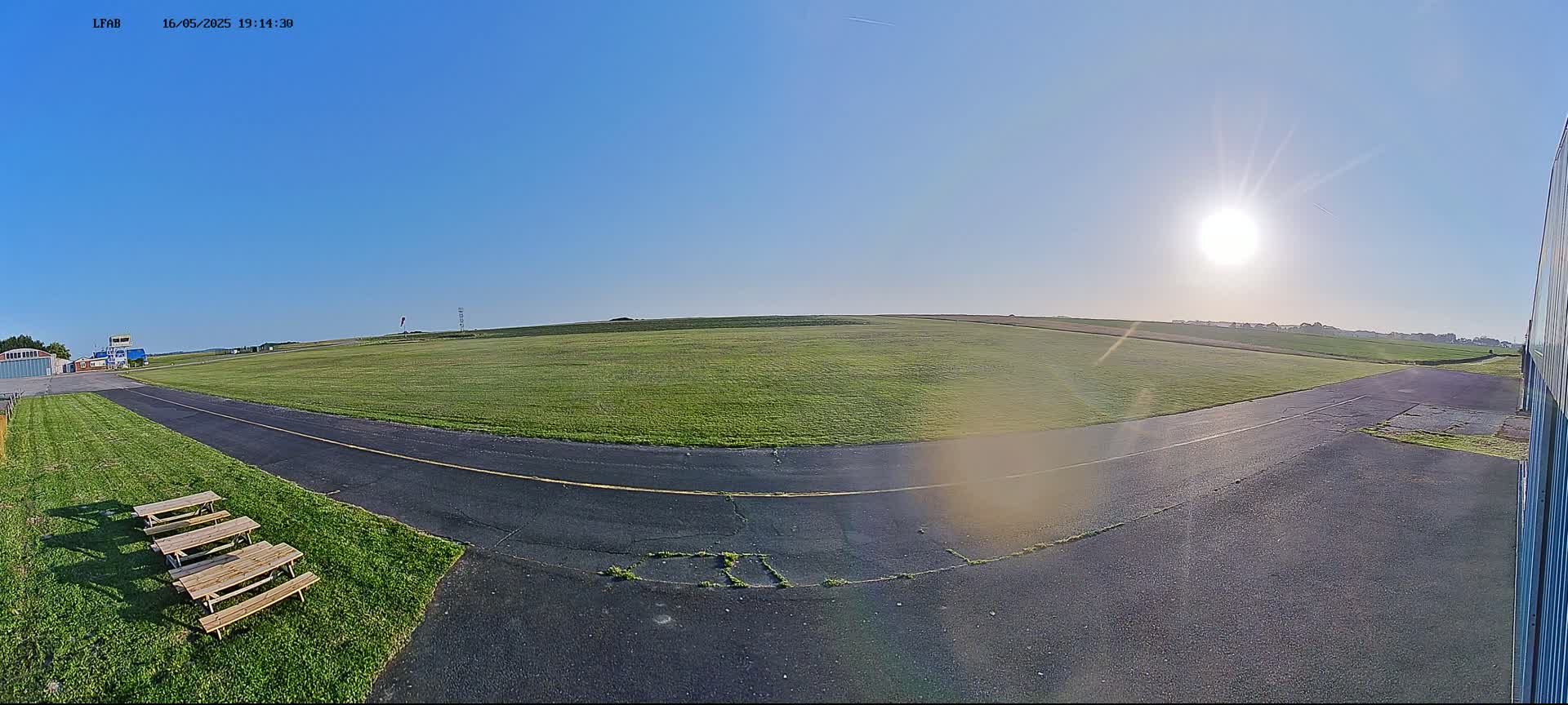A sunny day at a grassy airfield shows a paved runway and several picnic tables near a hangar.
