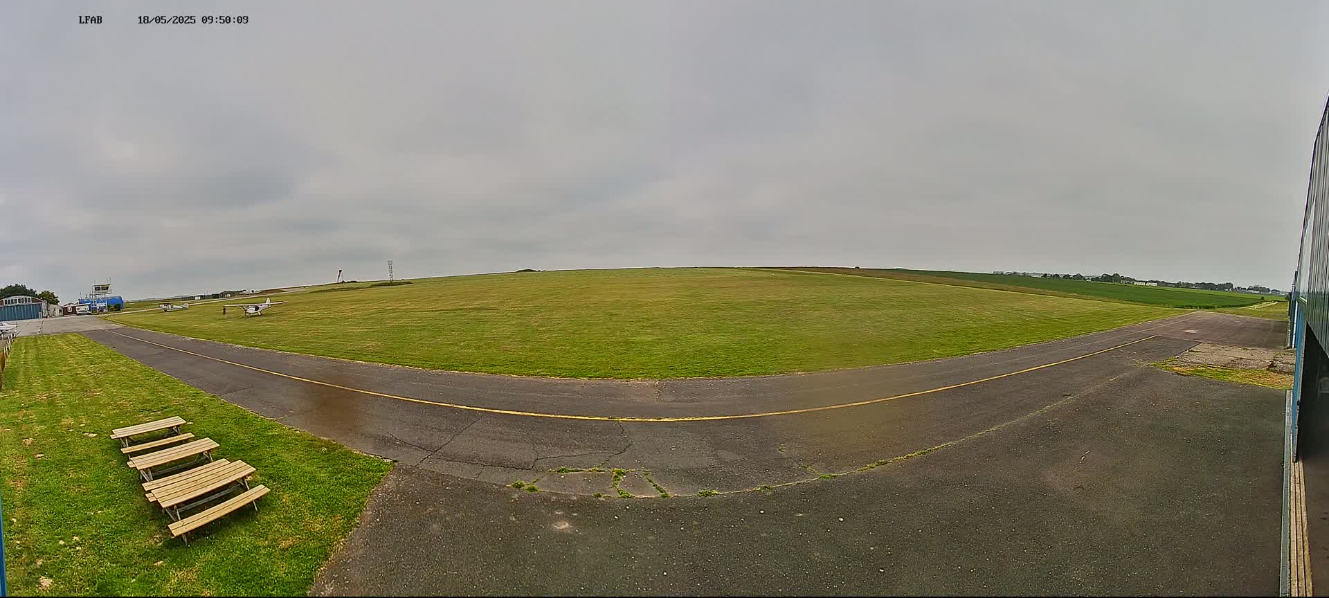 A small airfield with a grassy field, a paved runway, and several picnic tables sits under an overcast sky.