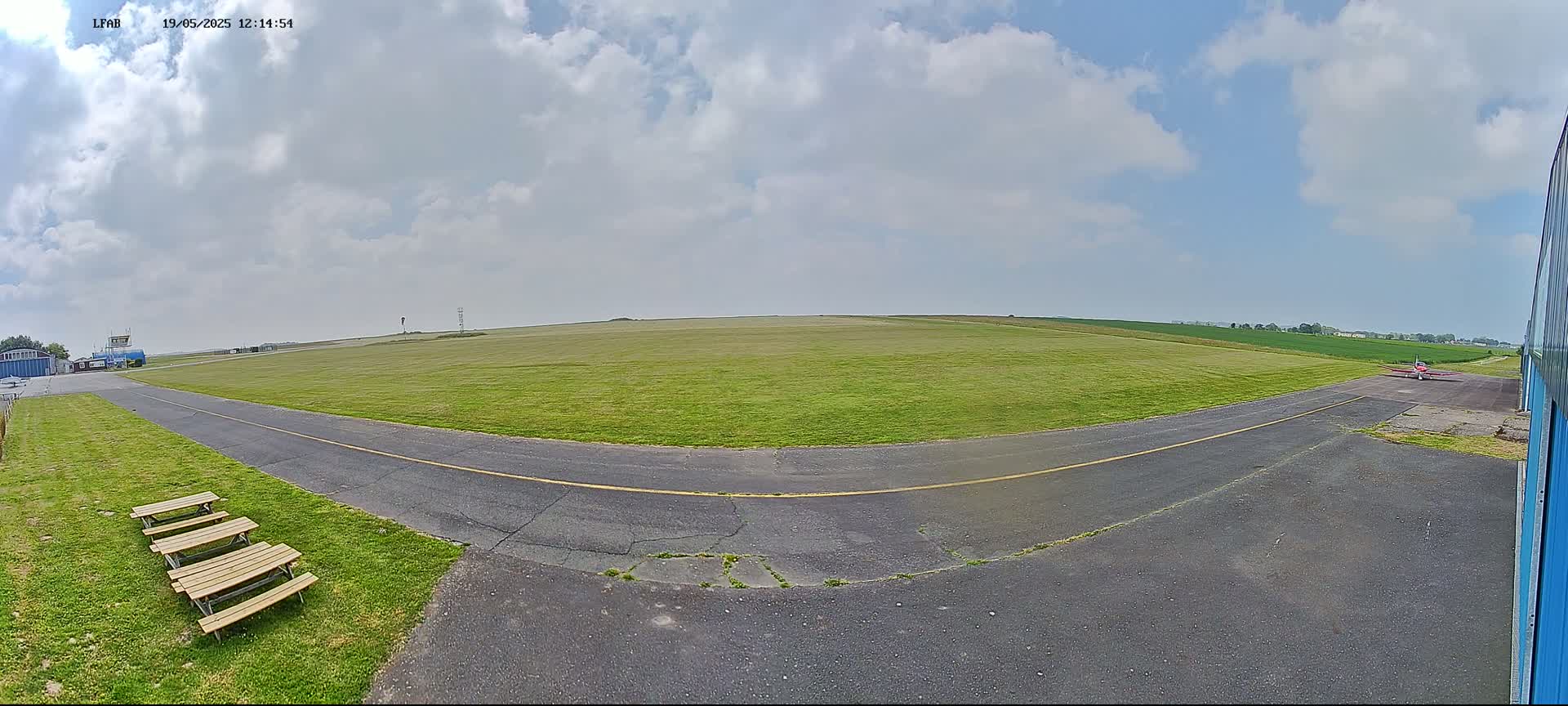 A small airfield with a grassy runway, a paved taxiway, and several picnic tables is visible under a partly cloudy sky.