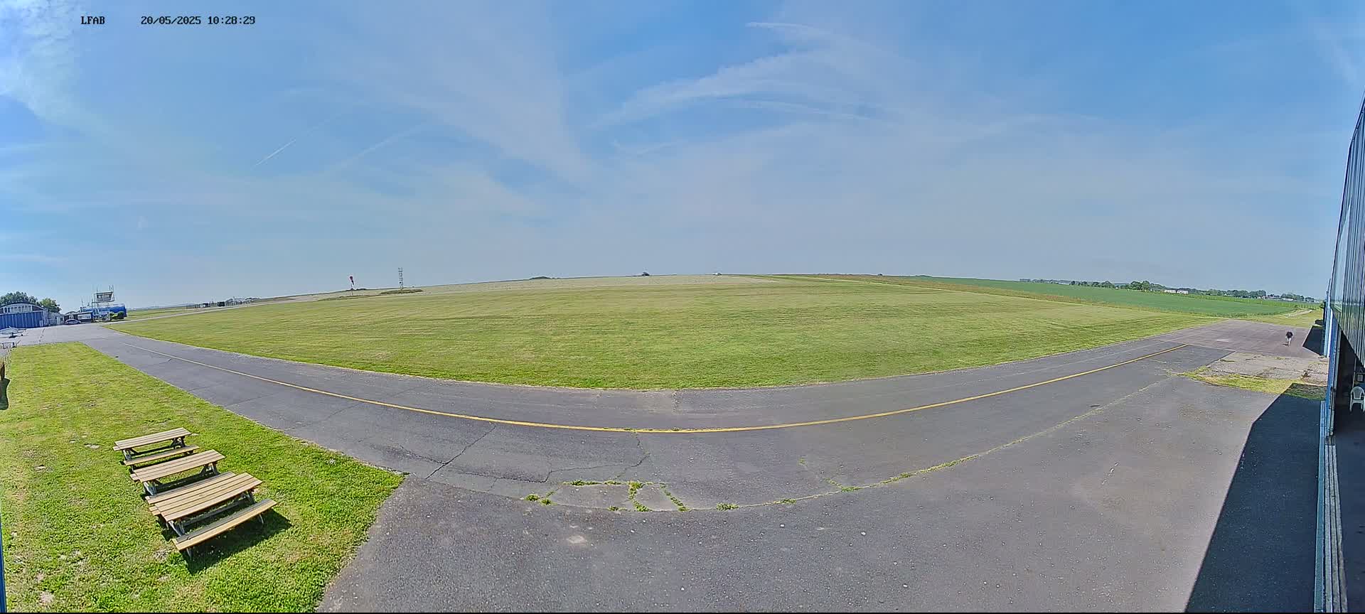 A grassy airfield under a mostly sunny sky with a paved taxiway curving toward a hangar, and picnic tables are visible in the foreground.