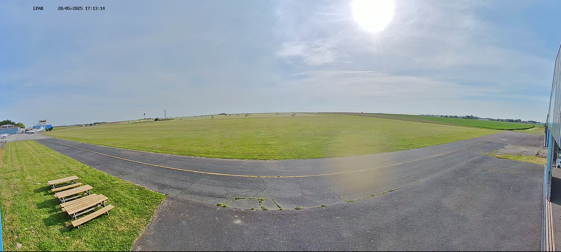 A sunny day at a grassy airfield shows a paved taxiway curving toward a large field of hay bales under a mostly clear sky.