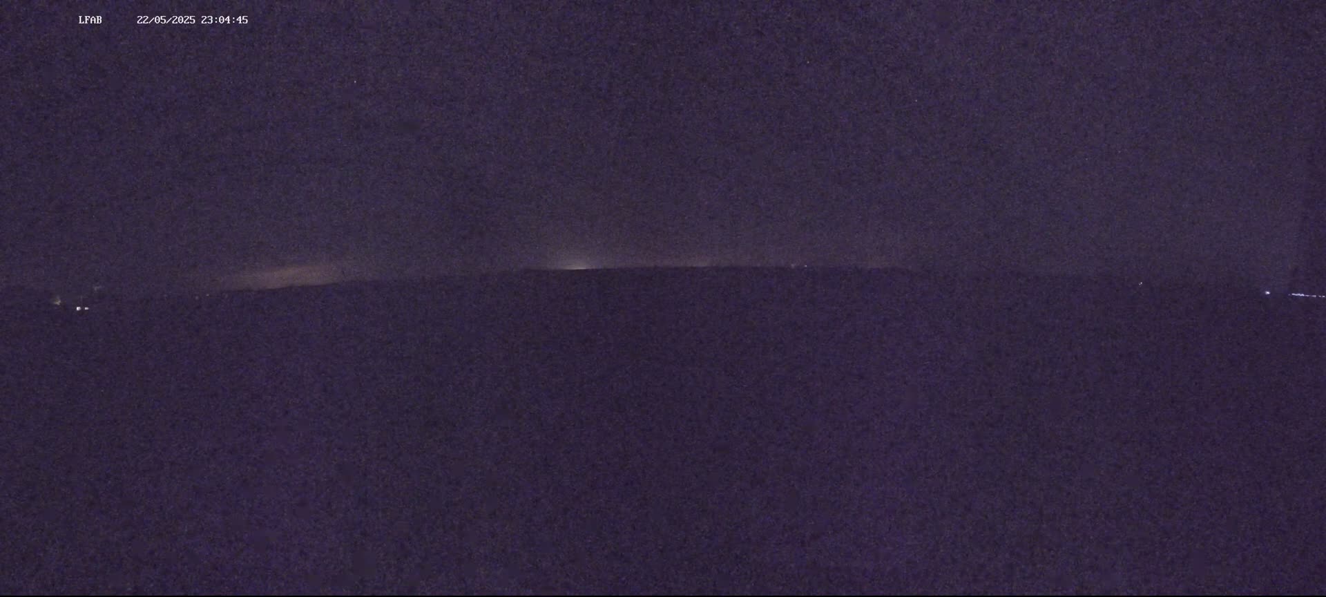 A dark, nighttime landscape shows a faint horizon line illuminated by distant lights under a mostly clear, starry sky.