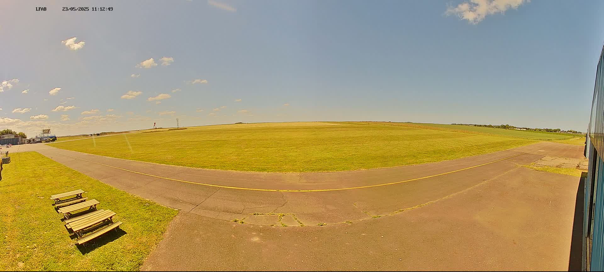 A mostly sunny day reveals a grassy airfield with a paved taxiway, picnic tables, and a hangar partially visible in the lower right corner.